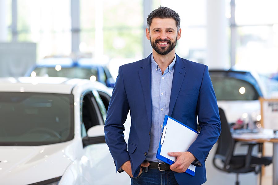 Business Insurance - Car Dealer With Dark Beard and Blue Suit Holds a Clipboard, Standing in Car Dealership Showroom