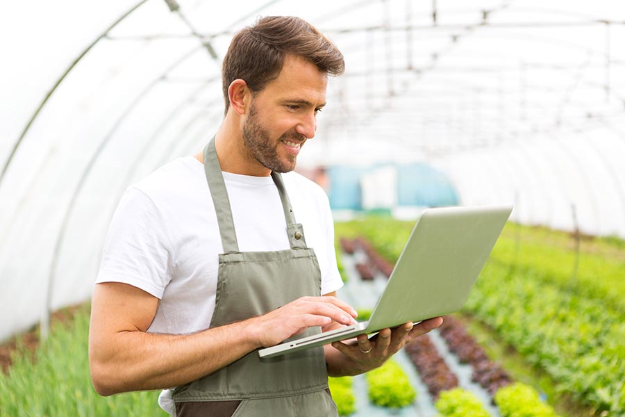 Contact Us - Young Business Owner Stands in a Greenhouse in an Apron, Using a Laptop and Smiling