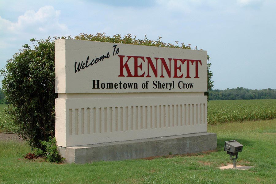 Kennett, MO Insurance - Large Tan Sign Reading Welcome to Kennett - Hometown of Sheryl Crow Sits in a Grassy Area, a Field Stretching Out Behind It