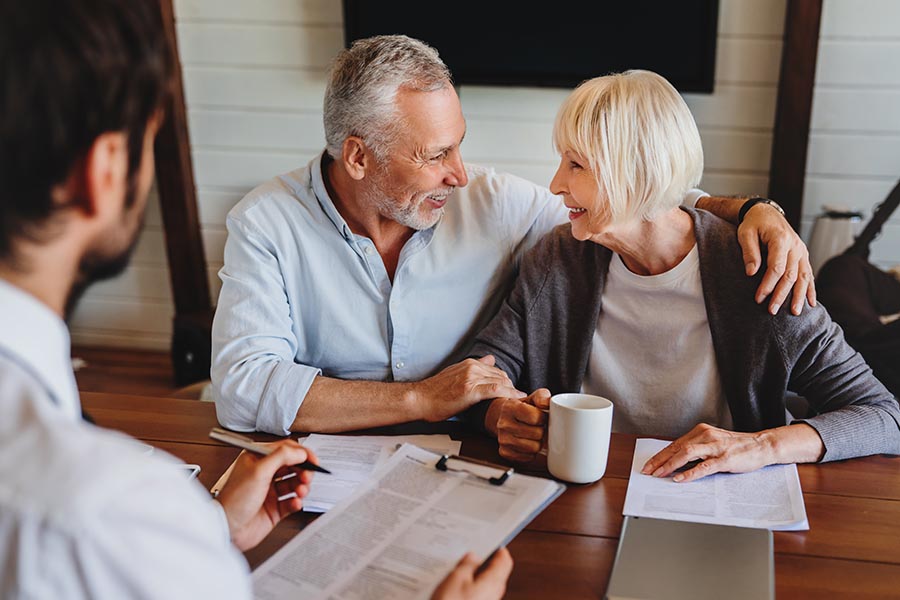 Securities & Investments - Senior Couple Looks Happy and Excited as They Review Paperwork With a Financial Advisor at His Desk, Wife Holding Coffee and Husband Wrapping Arm Around Her